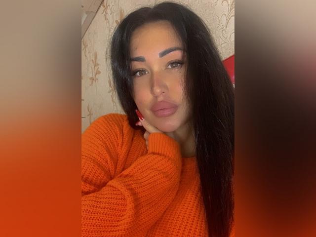 MelissaXCream met lang zwart haar, die een oranje gebreide trui draagt, haar volle lippen opgetrokken, haar kin rustend op haar hand, bruine ogen en rode nagels..