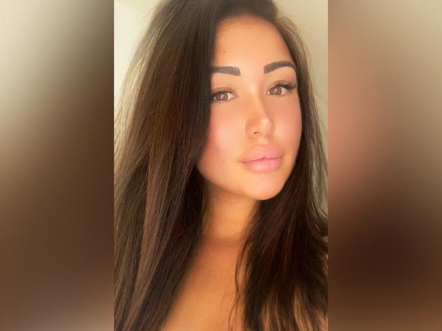 MelissaXCream hat langes, dunkelbraunes Haar, dicke Augenbrauen und pinken Lippenstift. Sie schaut mit einem leichten Lächeln in die Kamera. Ihre braunen Augen sind leicht nach oben geneigt..