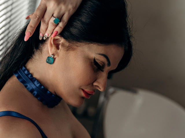 ClaireWarner with dark hair, blue choker, and matching earrings, has her head gently touched by a hand with pink nails and a teal ring, eyes closed, in a side profile.