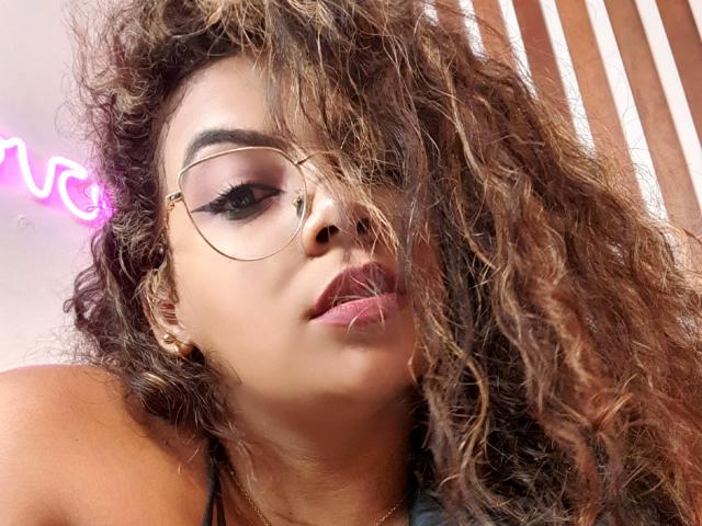 EbonyRose69 mit lockigem, braunem Haar, goldfarbenen Brillen und dunklem Eyeliner, zeigt volle, rosafarbene Lippen, leicht geöffnet, trägt ein goldenes Ohrring und blickt mit einem sexy, selbstbewussten Ausdruck nach oben..