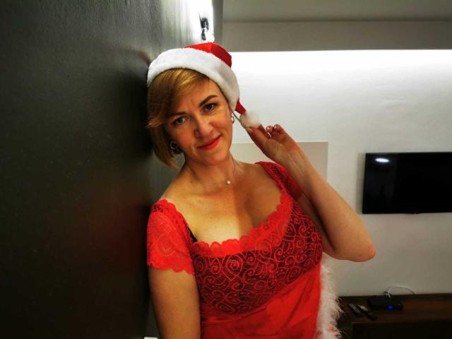 FelisiaStar with short blonde hair, red lace dress, and Santa hat, holding her hat, smiling, showing her tits, standing against a dark wall, looking cute.