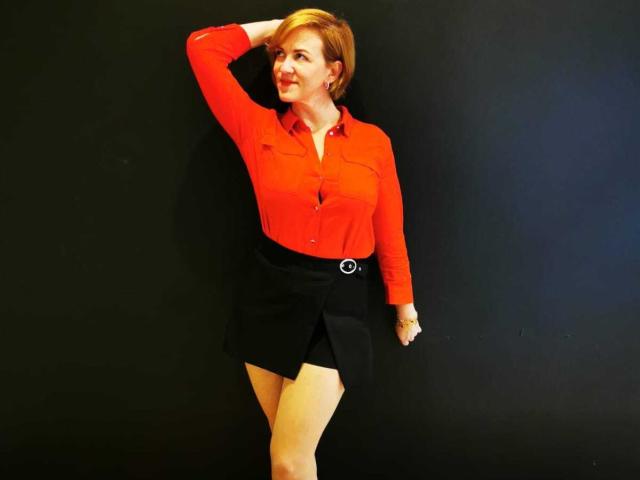 FeliziaStar with short red hair, red button-up shirt, black skirt with belt, standing against black wall, hand on head, smiling, showing off her tits and ass.