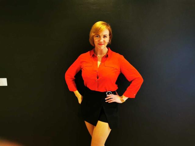 FelisiaStar stands against a dark wall, wearing a red shirt, black skirt with a belt, and yellow stockings. Hands on hips, smiling. Blonde, short hair, earrings.