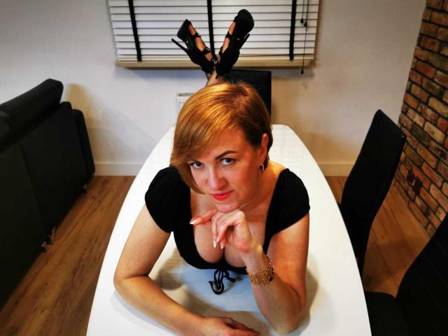 FeliziaStar with short red hair, black dress, and high heels, leaning on a white table, showing off her tits, with legs up, and a playful smile.