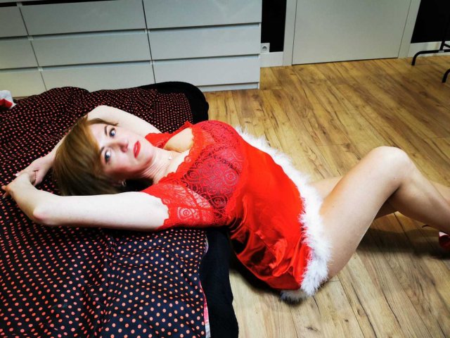 FeliziaStar is lying on a bed in a red lace lingerie dress with white fur trim, arms above her head, showing her tits and ass. She has red lipstick.