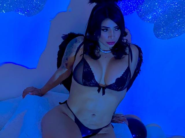 FransheskaParker with large tits, wearing black lace bra and panties, black feather wings, choker, tattoos on right arm, blue-lit, sitting on white fluffy surface, Brazzers watermark.