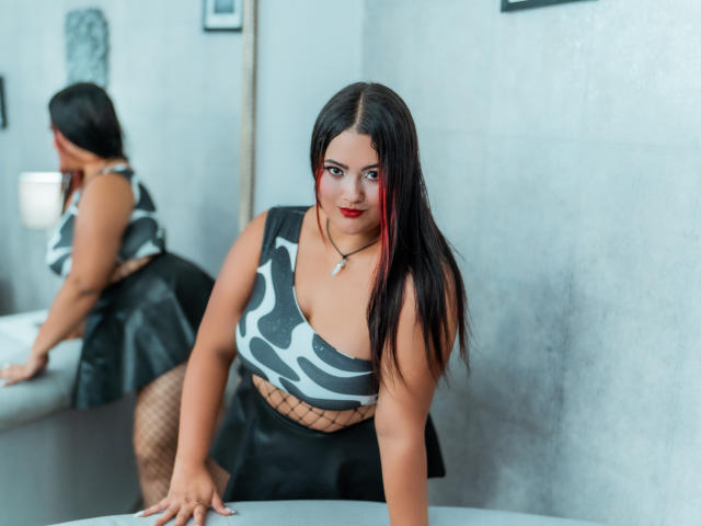 VaneParker a les cheveux foncés, du rouge à lèvres, et porte un haut gris à motifs avec découpes, une jupe noire, des bas résille, et elle est penchée en avant sur une table, avec un reflet dans un miroir..