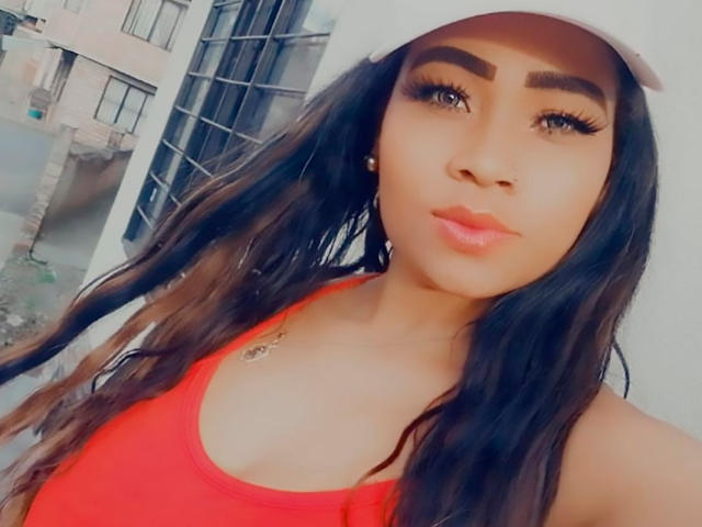 LinaaPrincess with long dark brown hair, thick eyebrows, big eyelashes, red lipstick, and a white cap, wearing a red tank top, showing her boobs.