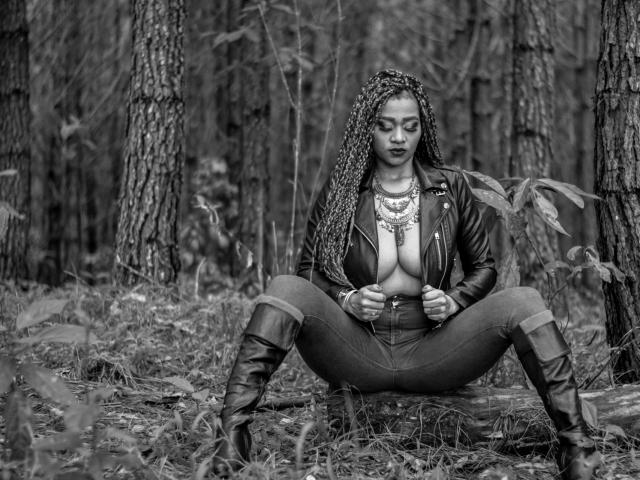 Black-and-white photo of LinaaPrincess sitting in forest, legs spread, wearing leather jacket, exposing her tits, braided hair, boots, and necklace, looking down with a serious expression.
