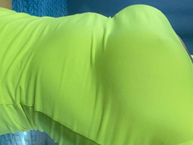 Misstyk's bright yellow, tight, form-fitting dress shows her curvy, round ass in the foreground, with smooth, slightly wrinkled fabric. Blue background is blurred.