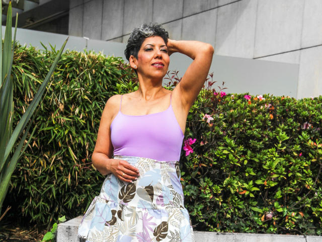 JuliaNewell has short curly black hair, a purple tank top, and a floral skirt, standing with one hand on her head, showing her tan skin and small tits.