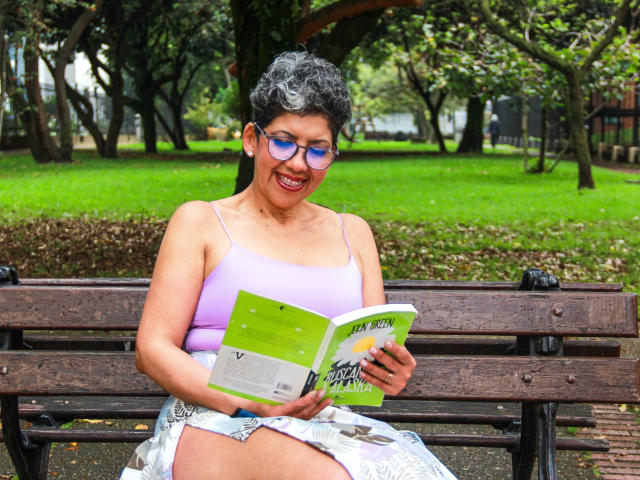 JuliaNewell with curly black hair, purple tank top, and glasses, sits on a wooden bench reading a green book, smiling with visible ass, in a park.