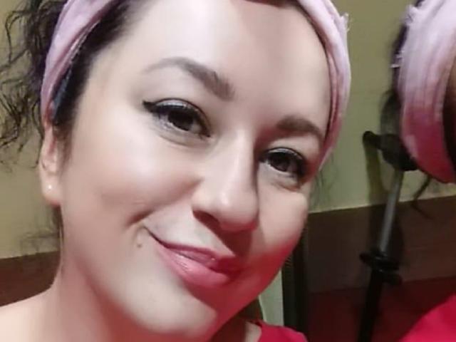 Tigra69I hat helle Haut, ein pinkes Stirnband, schwarzes, lockiges Haar, Eyeliner im Winged-Stil, pinken Lippenstift, einen kleinen Muttermal unter der Nase, lächelt leicht, ein rotes Hemd ist sichtbar, ein Teil des pinken Stirnbands ist auf der rechten Seite zu sehen..
