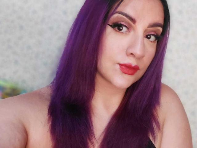AndreaSharick heeft paars haar, rode lippenstift en zwarte eyeliner. Ze kijkt in de camera met een lichte frons. Haar borsten zijn gedeeltelijk zichtbaar. Ze draagt een zwarte top..