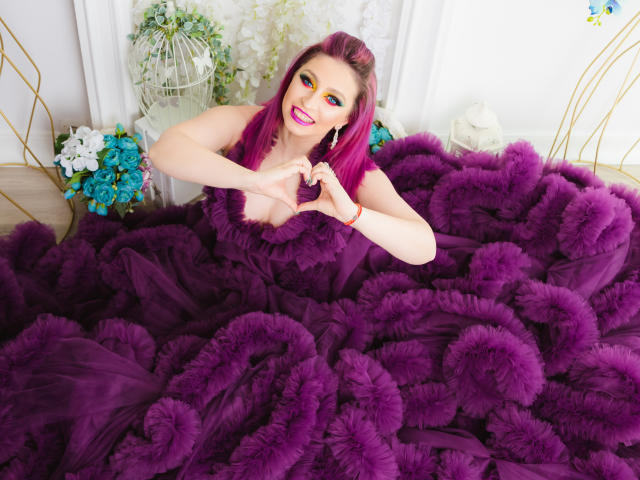 Agaa met roze haar, blauwe oogschaduw en een paarse jurk, die met haar handen een hartvorm maakt, omringd door een enorme, pluizige paarse rok. Borsten, mooi, lachend..