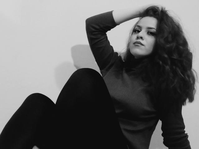 SpicyGrace, met krullend donker haar, gekleed in een zwarte coltrui en zwarte broek, zit met één arm achter haar hoofd, lichtelijk opzij kijkend, op een zwart-wit foto..