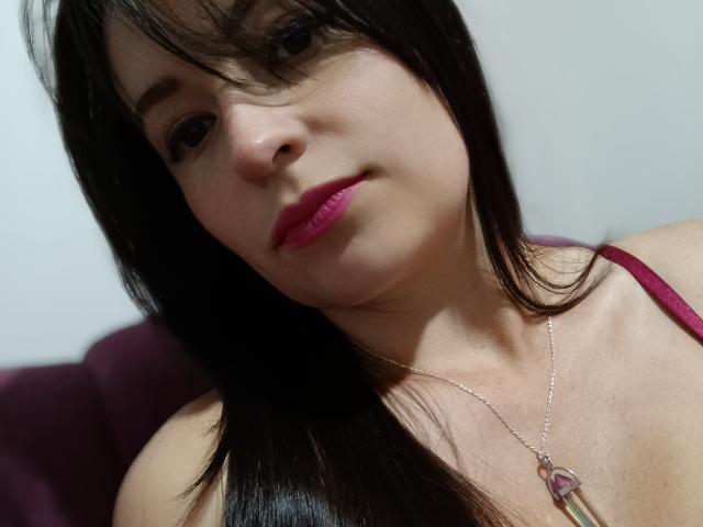 VaaleryStark with dark hair, pink lipstick, and a silver necklace with a heart-shaped pendant, wearing a red strap, looking at the camera with a slight smile.