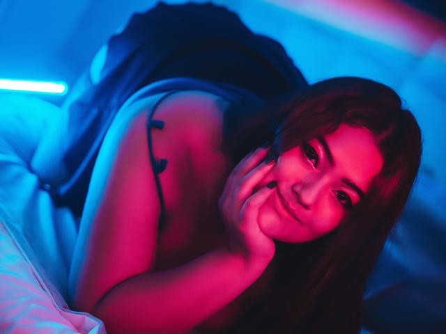 VaneParker ligt op een bed, op haar buik, waarbij haar borsten lichtelijk zichtbaar zijn. Ze draagt een zwarte top met bandjes en wordt verlicht door roze en blauwe neonlichten..