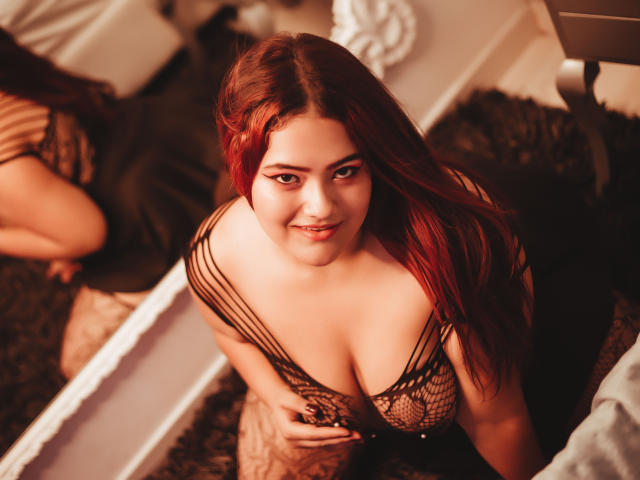 VaneParker with red hair, smiling down at the camera, wearing a black, netted top that exposes her large tits, kneeling on a dark rug, reflected in a mirror.