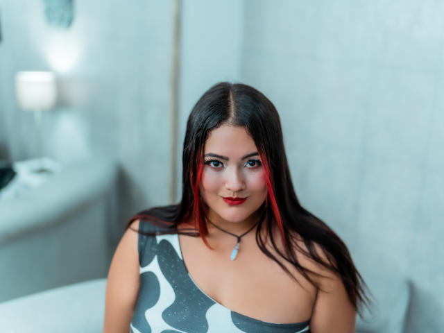 VaneParker tem cabelo castanho escuro, com mechas vermelhas, e veste uma blusa preta e branca com estampa, que deixa seus seios à mostra, com um pingente azul e batom vermelho, sentada em uma luz suave..