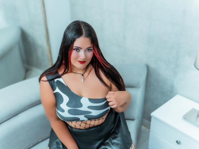 VaneParker con lunghi capelli neri, una mecca rossa, rossetto rosso, indossa una maglietta grigia e nera a motivi, calze a rete, una gonna in pelle nera, tiene in mano la maglietta, seduta su una sedia grigia, con una collana con una gemma..