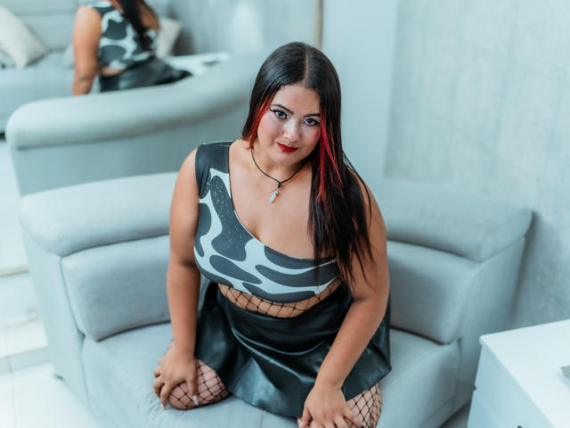 VaneParker, met zwart haar en een rode lok, en een gemiddelde borstmaat, draagt een zwart-wit topje, een zwarte rok, netkousen en rode lippenstift, en knielt op een grijze stoel..