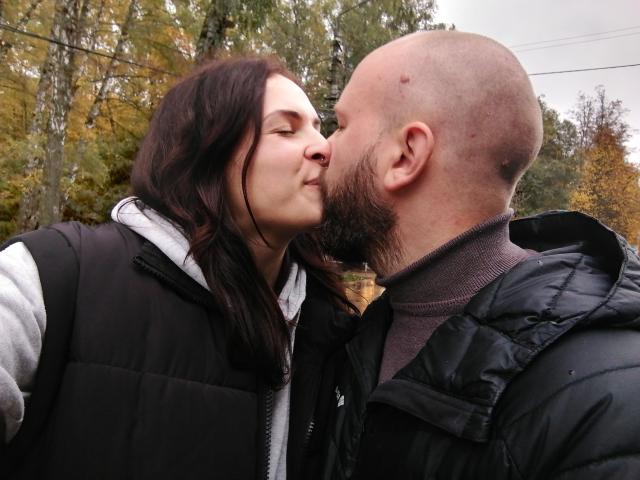 Una mujer con cabello oscuro besando a un hombre calvo con barba, ambos vistiendo chalecos negros y camisas grises, en un entorno al aire libre con árboles en el fondo..