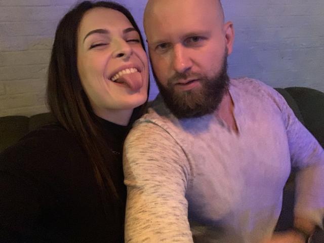 Uma mulher atraente com cabelos castanhos longos e seios, com a língua de fora, apoiada em outra mulher com a cabeça raspada e barba, vestindo uma camisa cinza, com uma expressão séria. Ambas sentadas em um sofá verde escuro..