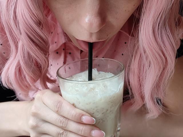 MissAstravert, avec ses cheveux roses ondulés, sirote un milkshake mousseux à travers une paille noire, sa main aux ongles roses tenant le verre, ses lèvres légèrement entrouvertes..