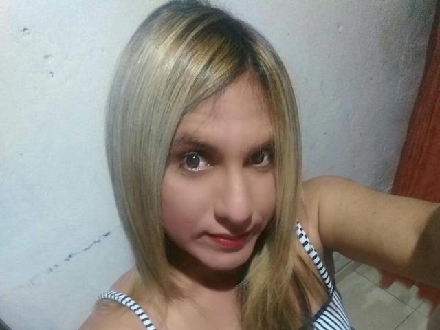 JaneStonesTs hat blondes Haar, helle Haut und trägt ein gestreiftes Tanktop. Auf dem Selfie trägt sie ein leichtes Lächeln und hat rote Lippen. Ihre Brüste sind klein und niedlich..