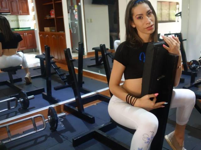 Allysxoxo, avec une poitrine de taille moyenne, assise sur un banc de musculation dans une salle de sport, portant un crop top noir et des leggings blancs, tenant une machine de musculation, son reflet se voyant dans un miroir..