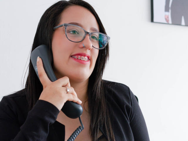 NatalieHarris with glasses, black shirt, holding gray phone to ear, smiling with pink lipstick, ring on finger, dark straight hair, white background, upper body, slightly angled.
