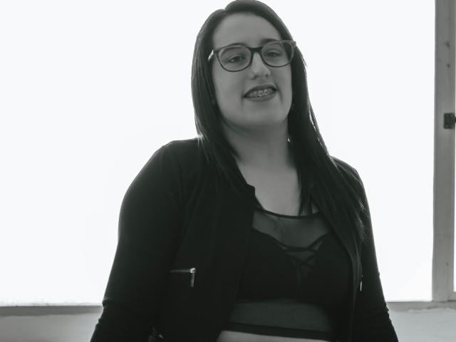 NatalieHarris has dark, straight hair, glasses, and is wearing a black cardigan over a sheer top, showing her tits. She's smiling with a slightly open mouth. Black-and-white photo.