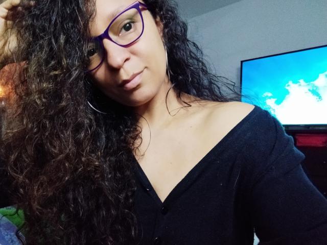 Angelonix with curly black hair, purple glasses, and a black off-shoulder top, smiling subtly, showing her tits, in a cozy room with a TV in the background.