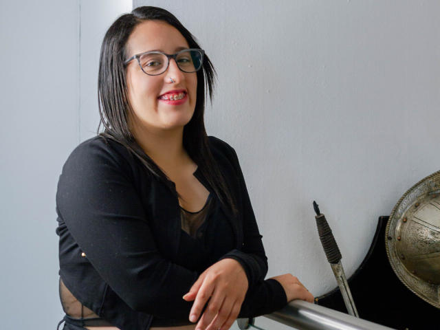 NatalieHarris with black hair, glasses, and red lipstick, wearing a black cardigan and black top, smiling at the camera, hands on a metal bar, pussy barely visible.