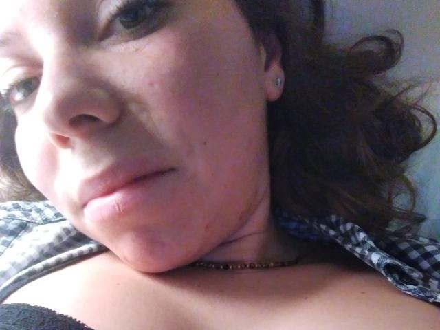Een selfie van Katarigna, waarop haar lichte huid, bruine golvende haren, kleine oorbellen, subtiele lipstick, een zwarte bh en een geruite blouse te zien zijn, met haar kin lichtjes gekanteld..