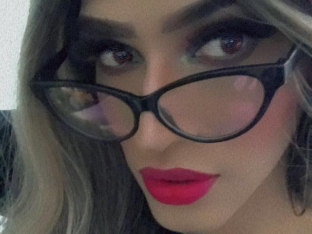 Cristal, con gafas de montura gruesa negra, lápiz labial rojo y cabello rubio, mira hacia arriba con ojos marrones. Su maquillaje es oscuro y ahumado, realzando su aspecto sensual..
