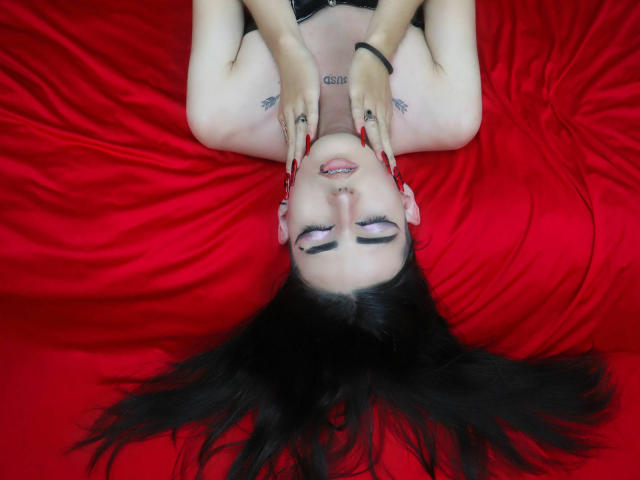 TokyoEvans, sdraiata su seta rossa, con le mani sul viso, i capelli neri sparsi, le unghie rosse, tatuaggi e un piercing al naso, che appare rilassata e sensuale..