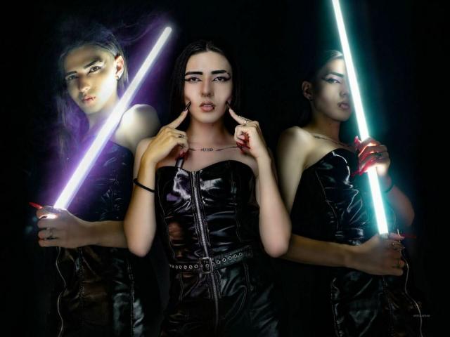 Tre TokyoEvans con capelli scuri, che indossano abiti di pelle nera lucida, e tengono luci al neon. La TokyoEvans al centro indica il viso, la TokyoEvans a destra ha una luce blu, la TokyoEvans a sinistra ha una luce viola..