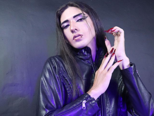 TokyoEvans, avec de longs cheveux noirs, une veste en cuir noir, du fard à paupières violet, du vernis à ongles rouge et un anneau au nez, regardant l'appareil photo, les mains près du visage, avec une lumière violette en arrière-plan..