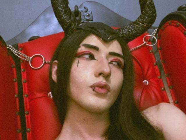 TokyoEvans con una máscara negra con cuernos, sombra de ojos roja, tatuaje de una cruz negra en la mejilla, labios fruncidos, cabello negro, silla de cuero rojo con anillos de metal, mirando hacia un lado..
