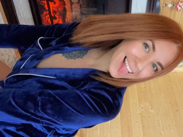 Misstyk with red hair, blue velvet robe, white earbuds, tongue out, smiling, tattoo on chest, wooden floor, fireplace in background. Boobs not visible, pussy not visible.