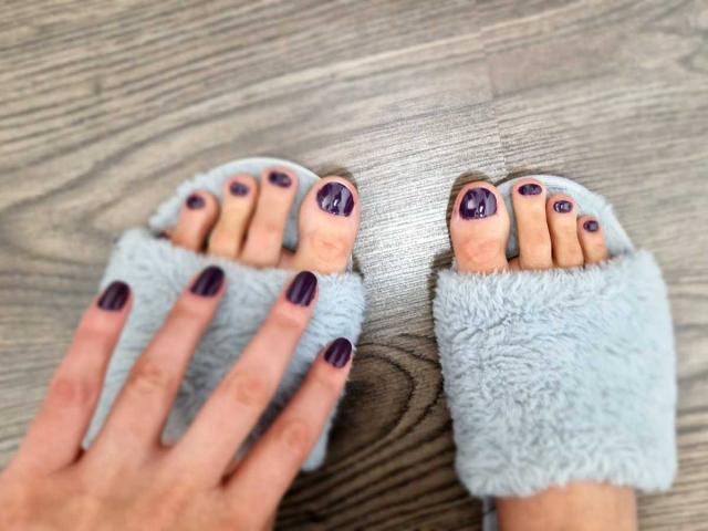 JoliDanielle's feet with purple toenails, wearing fluffy light blue socks, on a wooden floor. Her hand with matching purple nails rests beside her left foot.