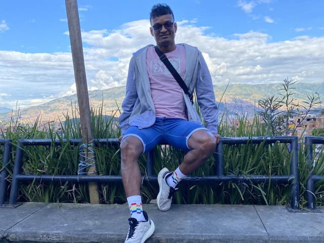 JacksonCooper sitting on a metal railing, wearing a pink tee, blue shorts, gray jacket, white socks with rainbow stripes, and white sneakers, with a black strap across chest.