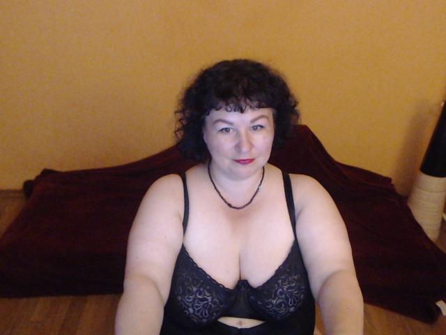 ErinSmart a les cheveux noirs bouclés, une peau pâle, et porte un soutien-gorge en dentelle noire, qui met en valeur sa poitrine généreuse, ainsi qu'un collier. Elle est assise sur un plancher en bois, recouverte d'une couverture rouge..