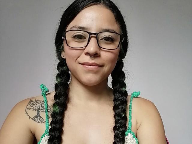SamyBrook, con trenzas negras, reflejos verdes, gafas, un piercing en la nariz y un tatuaje de un árbol en el hombro derecho, sonriendo y mostrando sus pechos con una blusa verde y blanca..