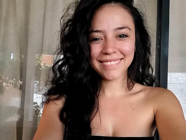 SamyBrook, avec ses cheveux noirs ondulés, sourit, montrant ses épaules dénudées et ses petites seins, debout devant une fenêtre avec des rideaux transparents. Elle a la peau claire et les yeux marrons..