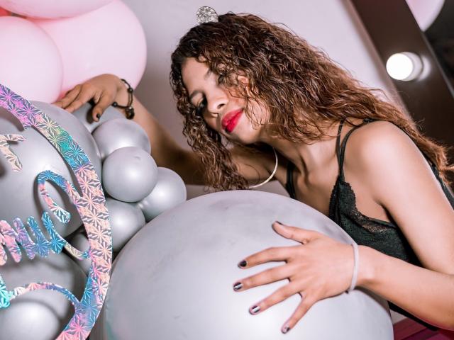 Ariana, uma mulher atraente com cabelo castanho encaracolado, batom vermelho, blusa preta de renda, tocando um grande balão prateado, com a mão sobre ele, usando uma pulseira, ao lado de balões rosa e um número decorativo..
