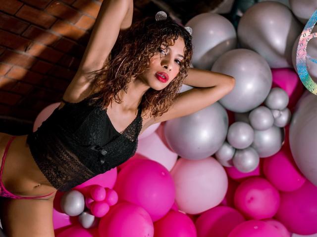 Ariana, una ragazza attraente con i capelli castani ricci, indossa una maglia nera in pizzo e biancheria intima rosa, appoggiata a un mucchio di palloncini rosa, grigi e argentati, con le braccia alzate e il rossetto rosso..