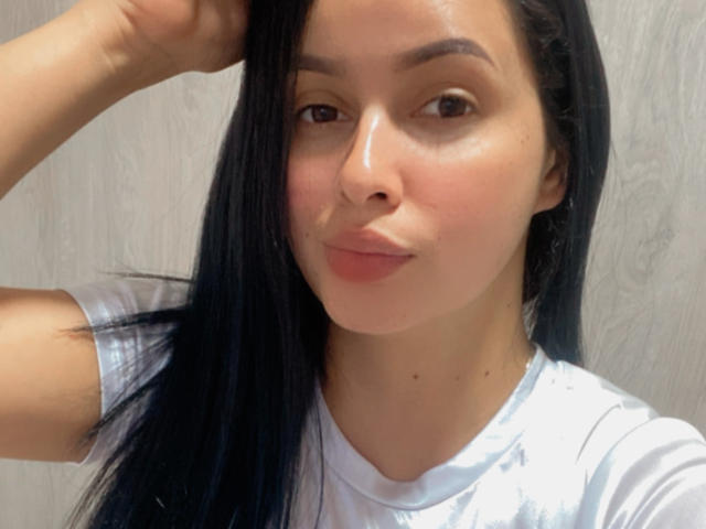 EmilyParkerr has long black hair, brown eyes, and a cute smile. She's wearing a white shirt, with a slight blush on her cheeks, looking at the camera.